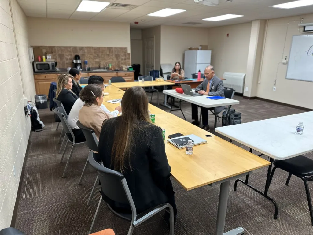 People sitting at a long L-shaped desk in a conference room with a trainer sitting across from them.
