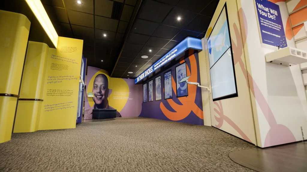 The inside of the People of Hope museum showing on the right four screens that visitors can view to learn more about people experiencing very difficult situations with a data wall next to it. On the left not clearly show is another exhibit. Straight ahead is a curved wall with the photo of a man smiling and unreadable text.