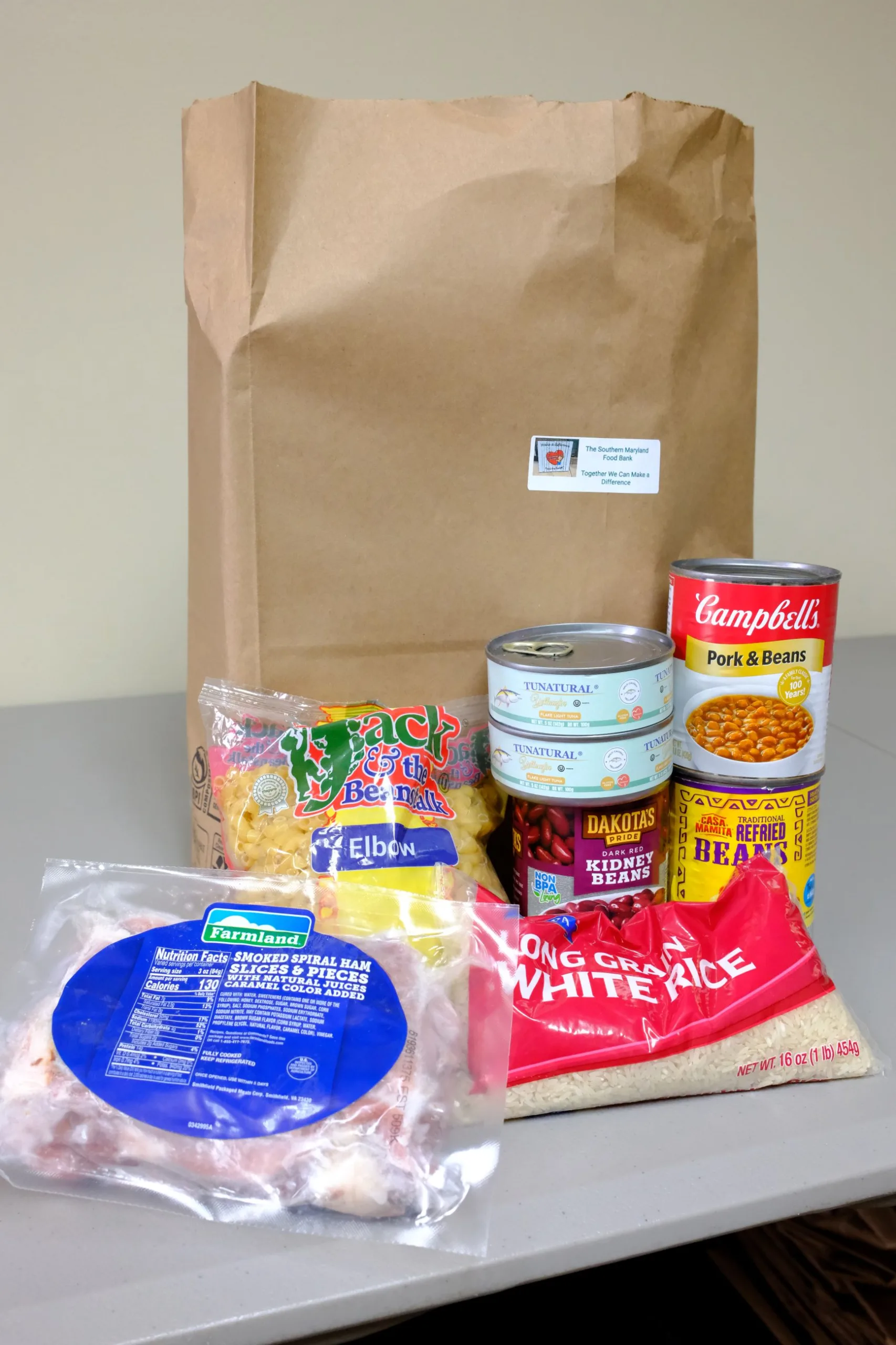 Large brown grocery bag with food pantry items in front of it including frozen meat, rice, pasta, beans, and tuna.